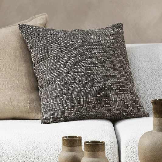 Alder cushion in dark chocolate with a light textured flect sitting on a grey sofa close up leaning against a beige linen cushion. Rust vases in the foreground