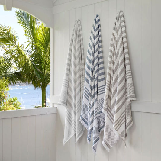 Striped beach towels called Bahama Peshtemal hanging up against a white wooden outdoor wall with the sea and palm trees in the background. 