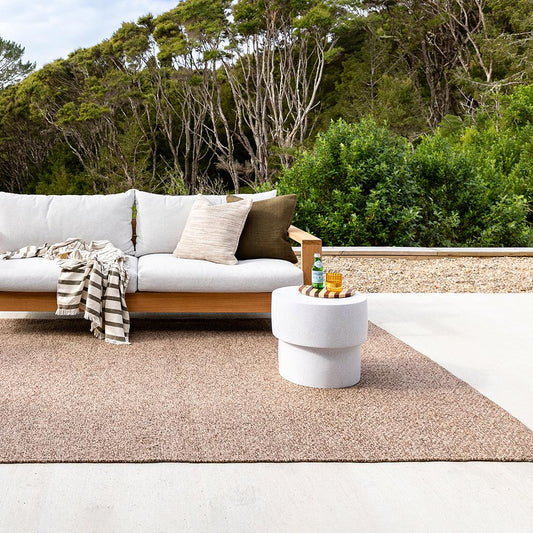 Burleigh Floor rug in teak in an outdoor setting with manuka bush in teh bank ground with a cream and wood sofa with outdoor cushions and a white concrete coffee table