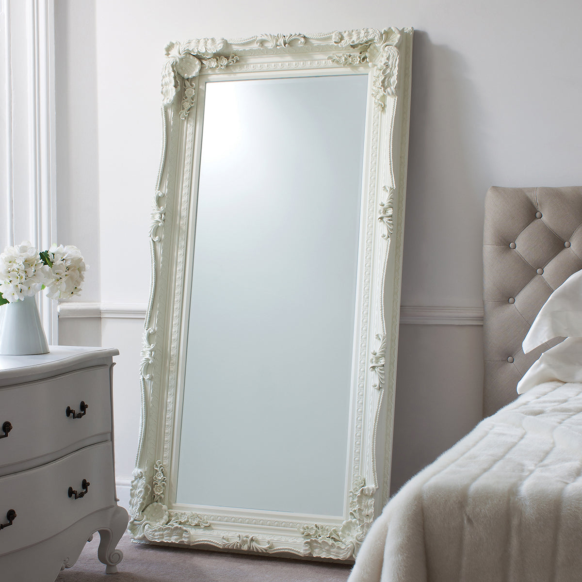 Carved ornate large white mirror leaning on a bedroom wall witih a luxurious white bed with uphostered headboard and french style dresser