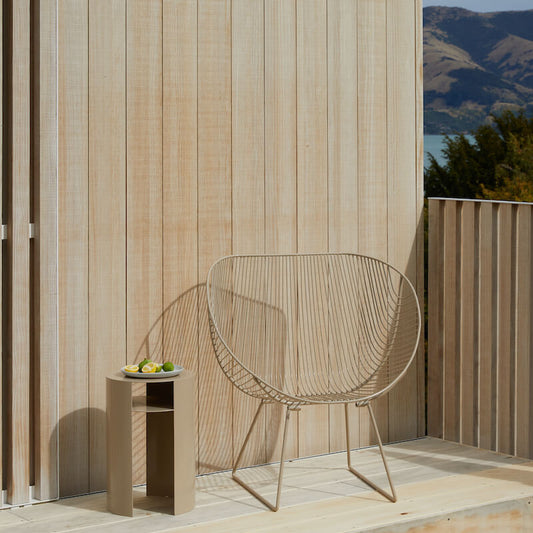 A putty coloured rounded wire chair called the Coroandel sitting on a wooden deck against a light wooden wall