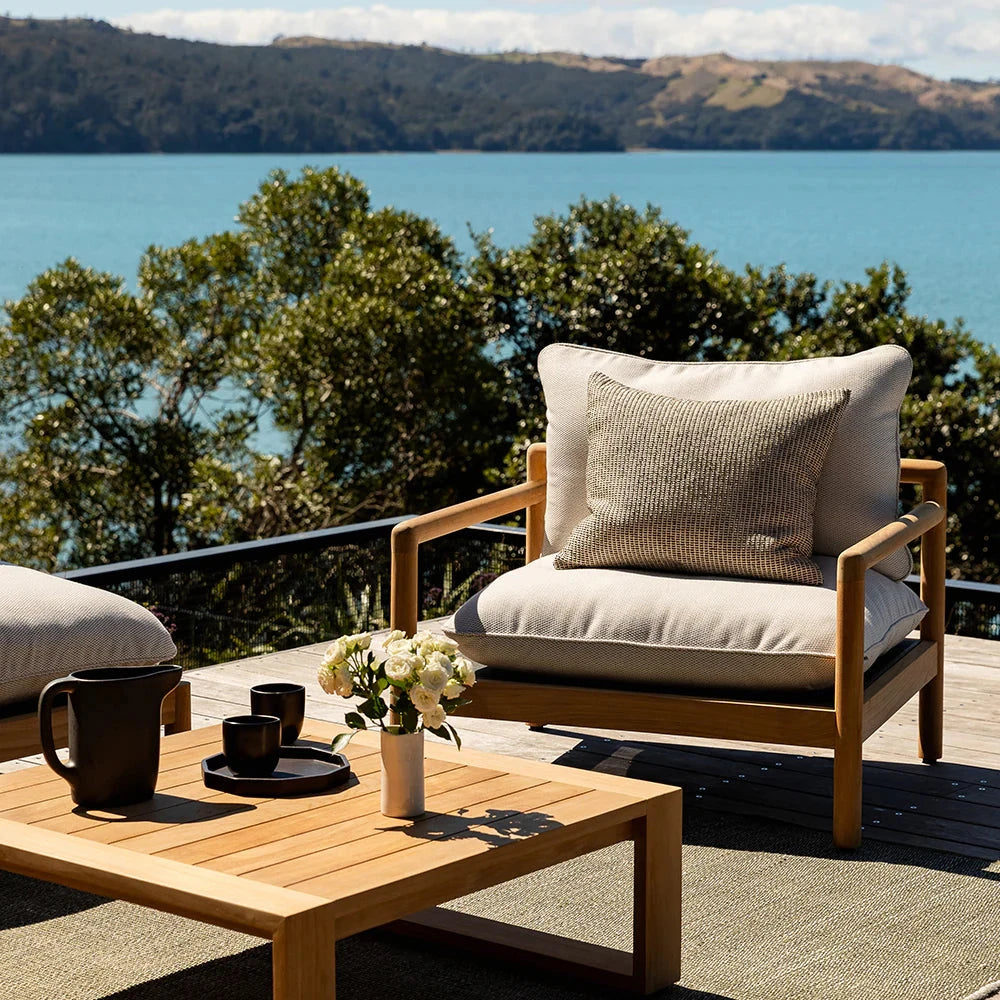 textured woven brown cushion on a cream and wooden outdoor chair on a deck with a lake and mountains in the background
