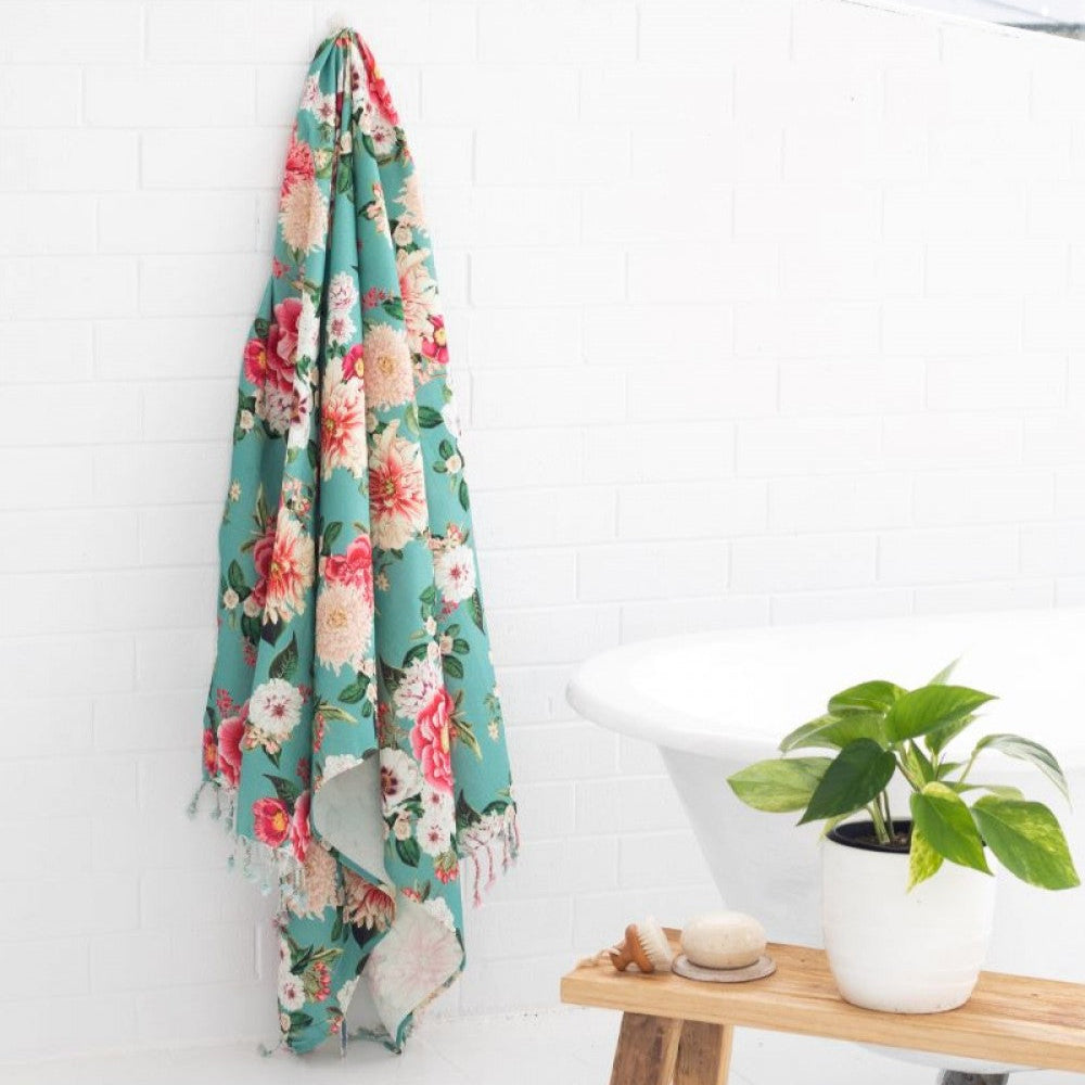 Fleur Turkish Towel, Aqua coloured towel with a bright large floral design and tassels, hanging on a hook in the bathroom