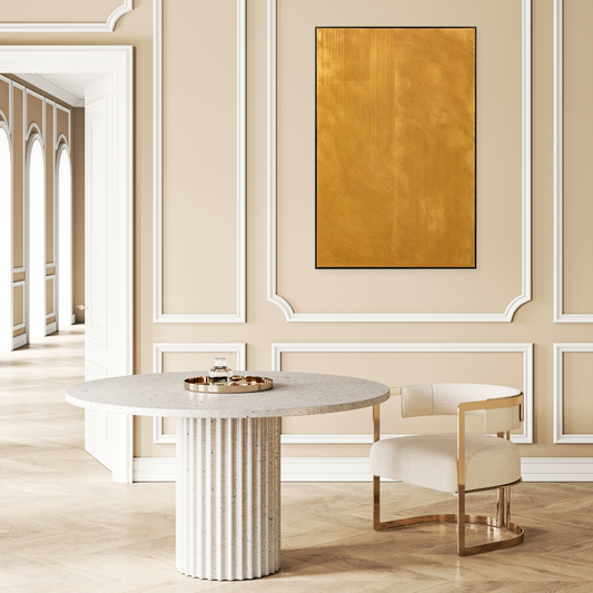 gold textured oil painting on a beige wall with panelling. Infront is a white marble round table and a gold and white fabric mid century style chair, both sitting on a pale wooden parquet floor
