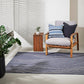 Blue rug with ivory swirly lines mimicking the river in a room with grey walls and flor, a wooden seat with great cushion and blue striped cushions and a ot with greenery and a coffee table made from a wooden pallet with a plant on top