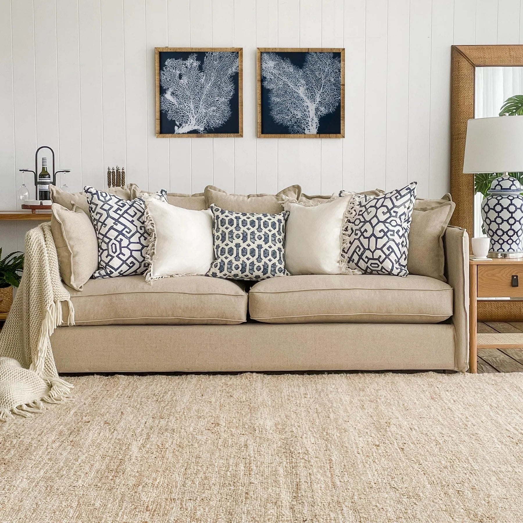 hamptons inspired lounge with coral images on navy background wall art. A beige cushion has lots of cream and blue and white cushions on with a wooden table and mirror to the side featuring a tall blue and white lamp
