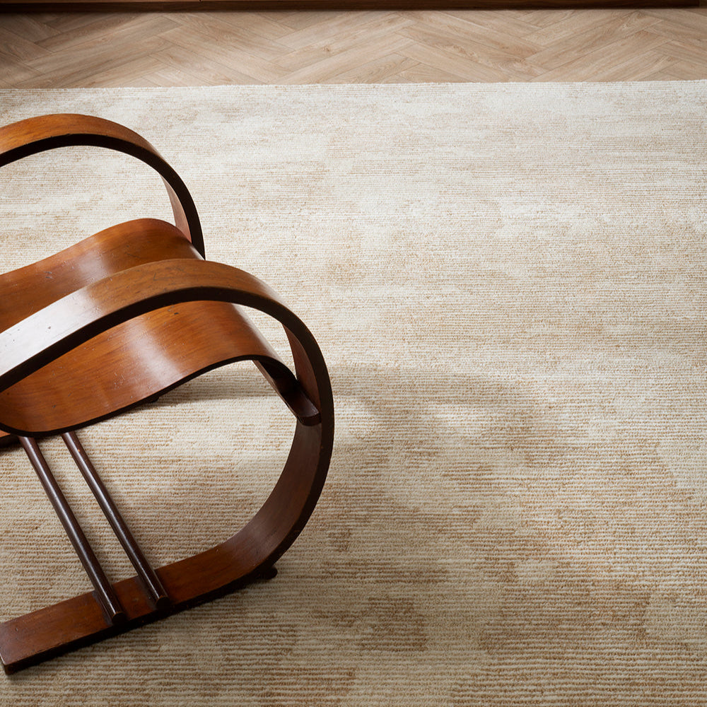 Beckett Floor rug a mottled beige rug with a wooden rocking chair sitting on it and a herringbone wooden floor
