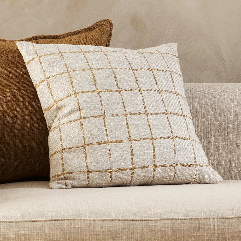 plain cream cushion with a ragged grid pattern design in ochre sitting on a cream sofa against a tobacco coloured plain cushion
