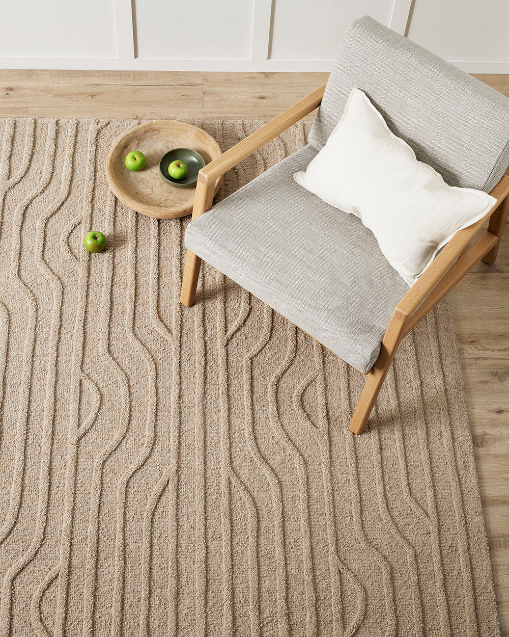 textured lines on a pebble coloured rug sitting on a wooden floor. A grey mid century style chair sits on the rug beside a wooden bowl of green apples