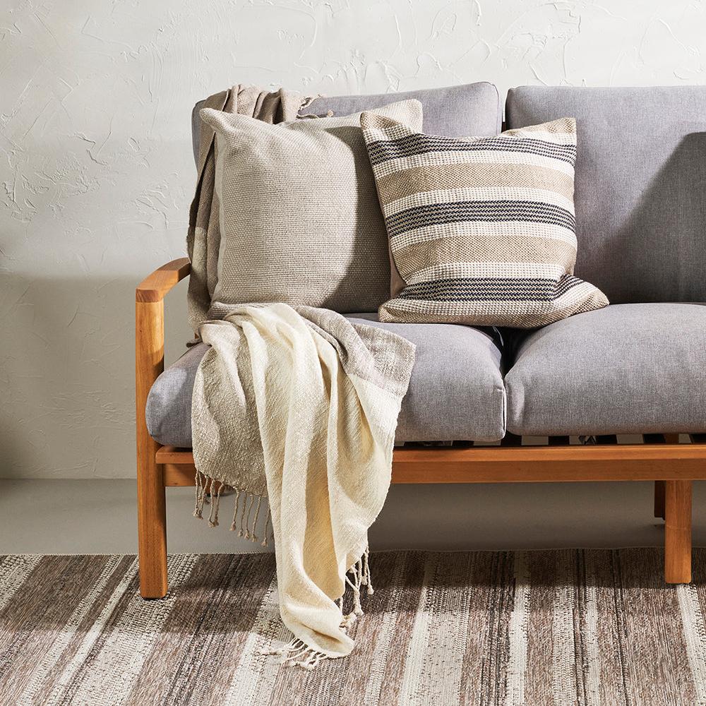 two cushions in neutral colours one striped and one plain on a grey sofa with wooden handles. 