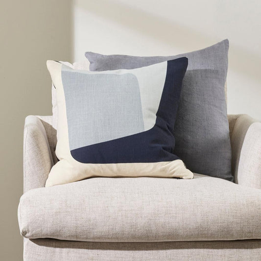 Maria cushion in a colour block patter of light blue, navy and pale blue sitting against a grey cushion on a linen beige chair