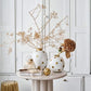 a grouping of round white vases with gold dots on a white marble side table that features other things like books, baubles etc. The vases have dried flower arrangements like hydrangeas and the room back ground is white with old fashioned edwardian shutters
